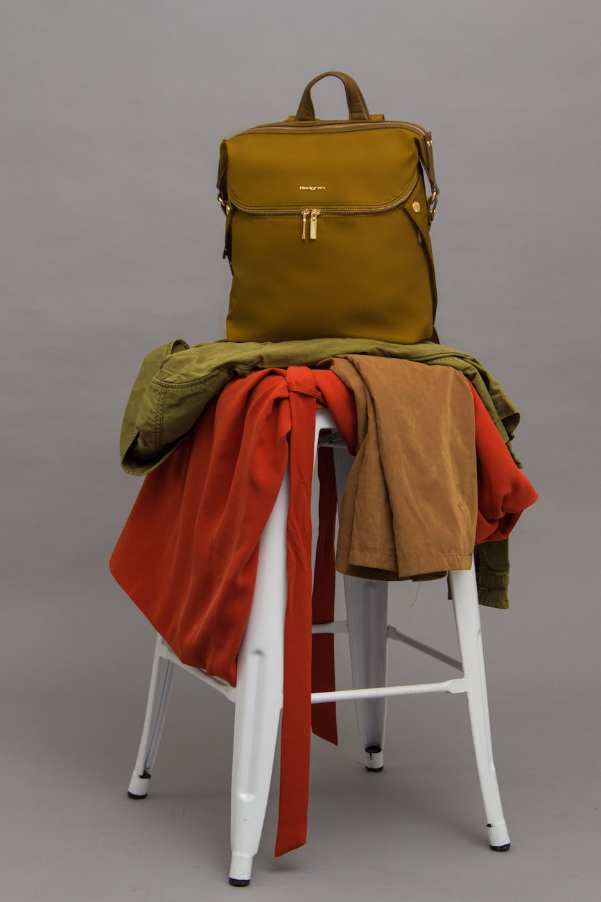 A fashionable olive green backpack placed on a white stool with vibrant clothes draped over it.
