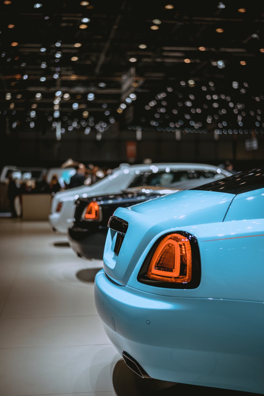 Rear view of luxury cars lined up at an indoor auto show with bright lighting.